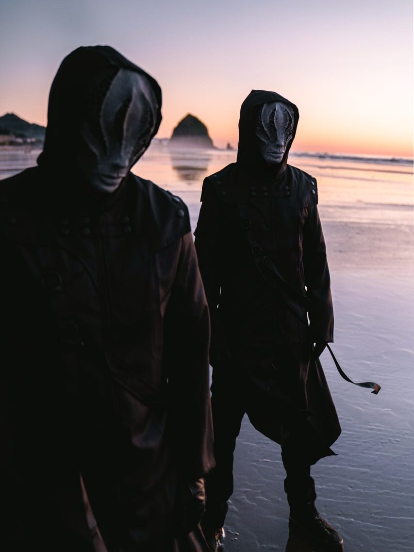 The two members of ABANDONED in masks and black coats on a beach at sunset